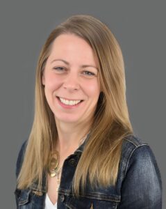 Smiling person with long hair wearing a denim jacket against a gray background