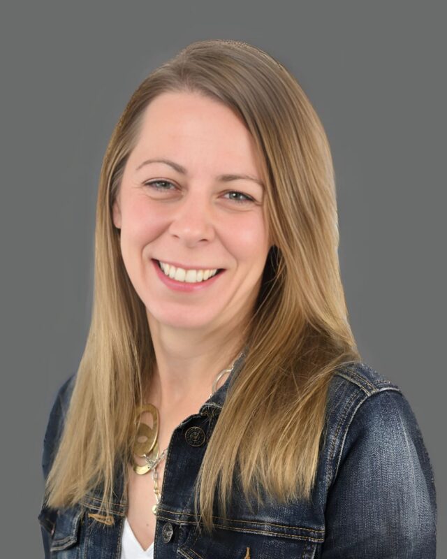 Smiling person with long hair wearing a denim jacket against a gray background