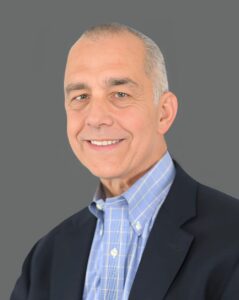 Smiling man in a suit jacket and blue shirt against a gray background.