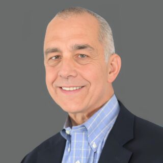 Smiling man in a suit jacket and blue shirt against a gray background.