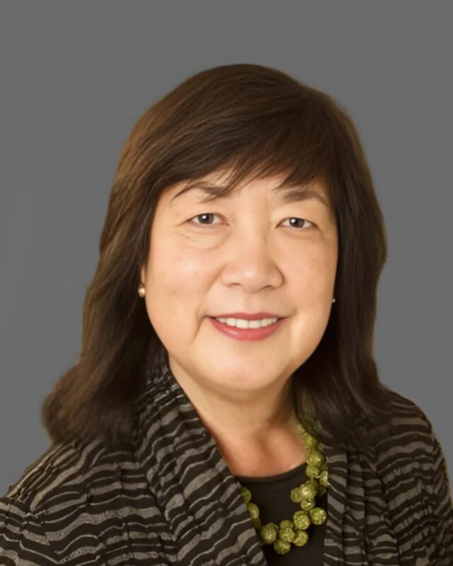 A person with shoulder-length hair and a patterned jacket smiling against a plain background.