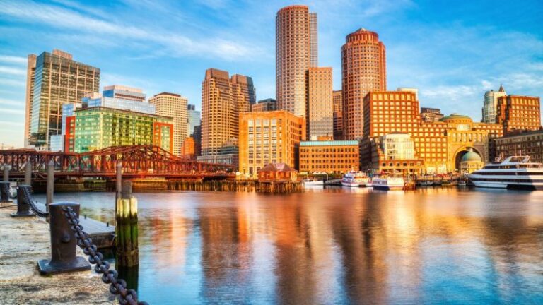 Skyline of a city with tall buildings reflecting on the water at sunset, under a clear blue sky.