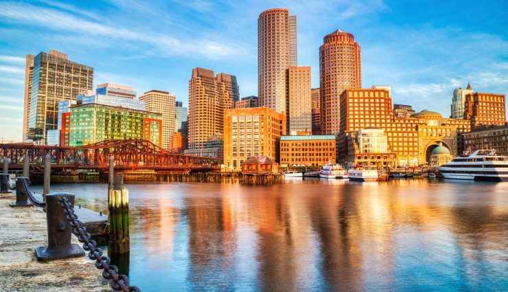 Skyline of a city with tall buildings reflecting on the water at sunset, under a clear blue sky.