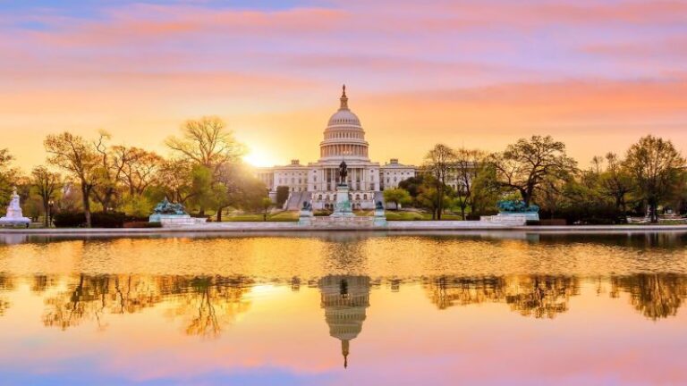U.S. Capitol building at sunrise with a vibrant sky and reflection in the water, surrounded by trees and statues.