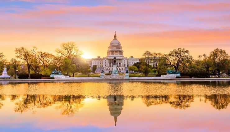 U.S. Capitol building at sunrise with a vibrant sky and reflection in the water, surrounded by trees and statues.