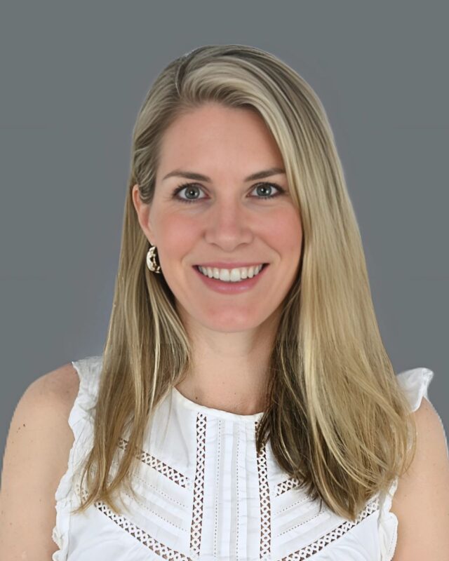 Smiling individual with long blonde hair wearing a white top, set against a plain gray background.