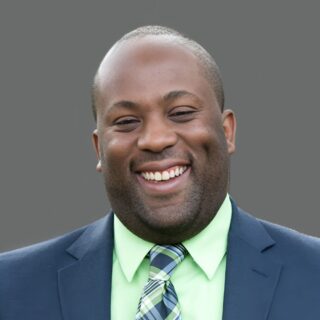 Smiling man in a suit, green shirt, plaid tie, against a gray background.