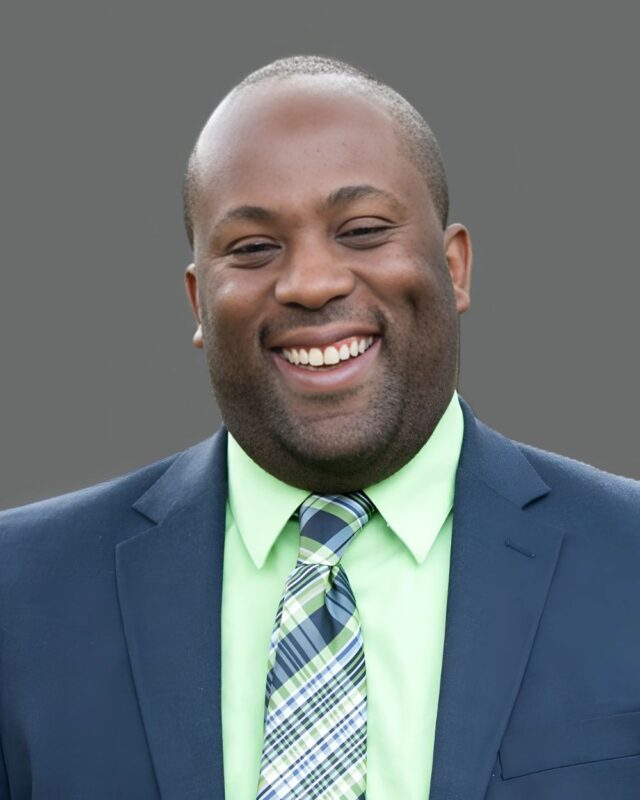 Smiling man in a suit, green shirt, plaid tie, against a gray background.