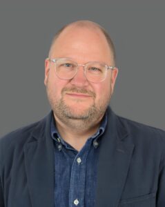 A person with glasses, wearing a dark jacket and blue shirt, smiling against a gray background.
