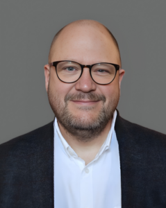 Smiling man with glasses, wearing a white shirt and dark jacket against a gray background