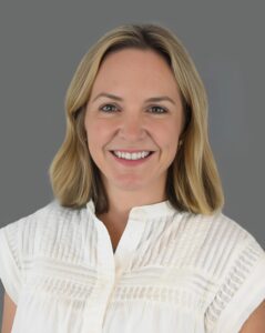 Smiling person with shoulder-length hair wearing a white shirt against a gray background.