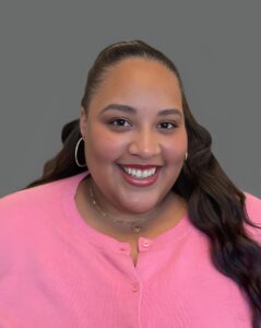 Smiling person wearing a pink top and hoop earrings against a gray background.