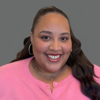 Smiling person wearing a pink top and hoop earrings against a gray background.
