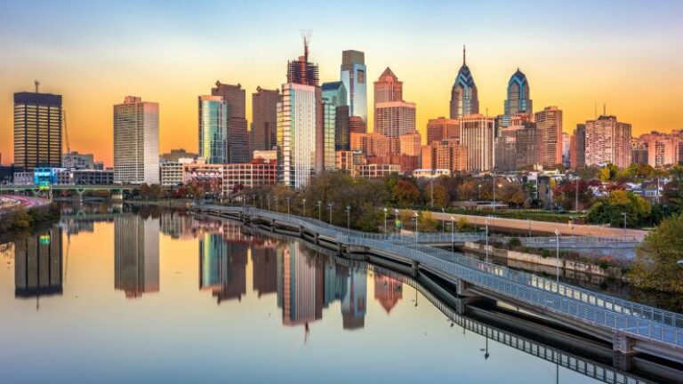 Philadelphia skyline at sunrise with skyscrapers reflecting in the calm river and a partially visible bridge in the foreground