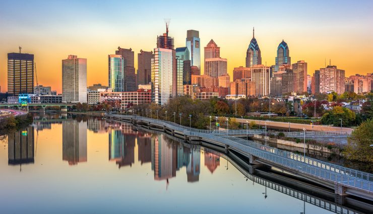 Philadelphia skyline at sunrise with skyscrapers reflecting in the calm river and a partially visible bridge in the foreground