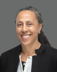Smiling person with braided hair, wearing a black jacket and white shirt, against a gray background.