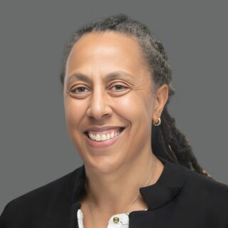 Smiling person with braided hair, wearing a black jacket and white shirt, against a gray background.