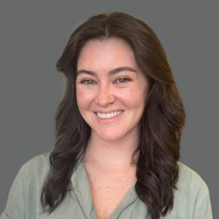 Smiling person with long dark hair and a nose ring wearing a green shirt against a gray background.