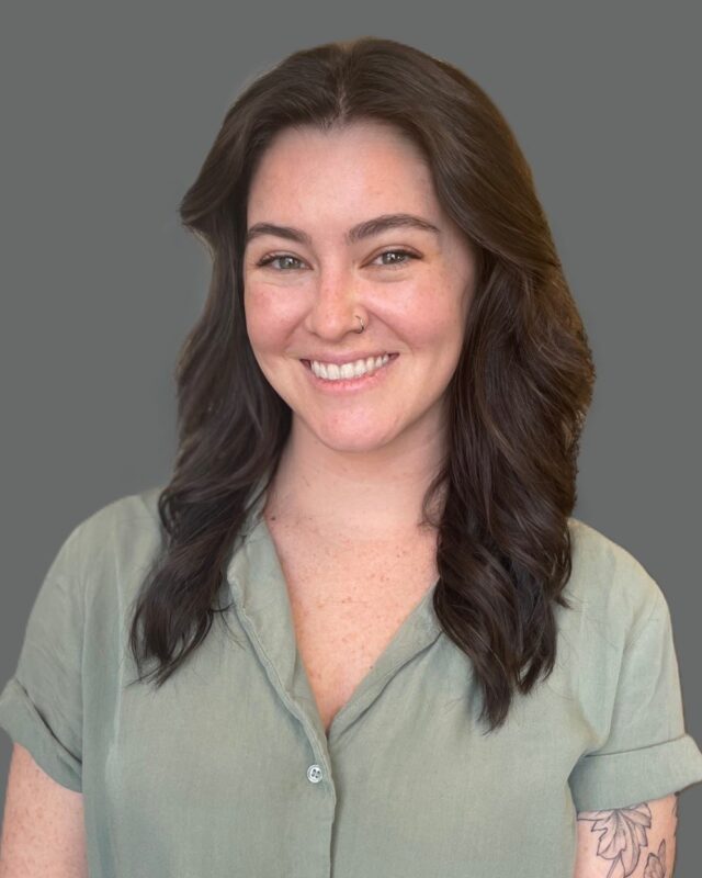 Smiling person with long dark hair and a nose ring wearing a green shirt against a gray background.