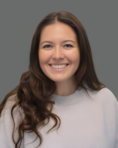 Smiling person with long brown hair wearing a light gray sweater against a gray background.