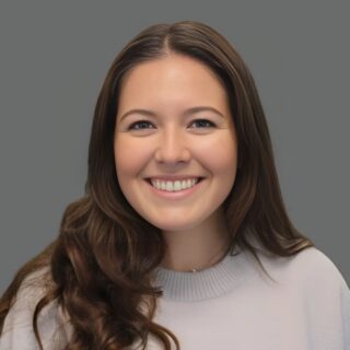 Smiling person with long brown hair wearing a light gray sweater against a gray background.