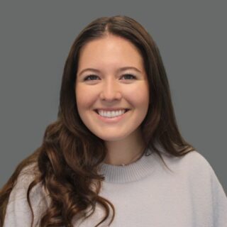 Smiling woman with long brown hair, wearing a light gray sweater against a plain gray background.