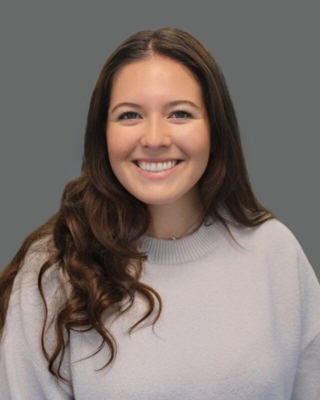 Smiling woman with long brown hair, wearing a light gray sweater against a plain gray background.