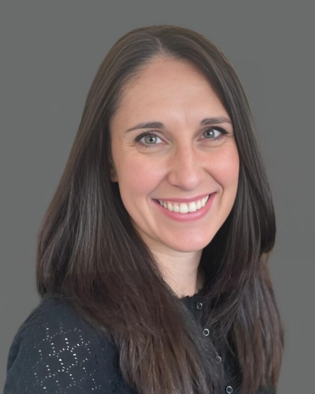 Smiling woman with long dark hair wearing a black top against a gray background