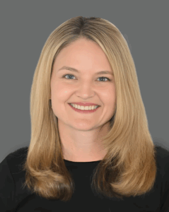 Smiling person with long blonde hair wearing a black top against a gray background.