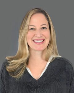 Smiling person with long blond hair wearing a gray blouse against a gray background.