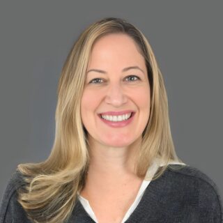 Smiling person with long blond hair wearing a gray blouse against a gray background.