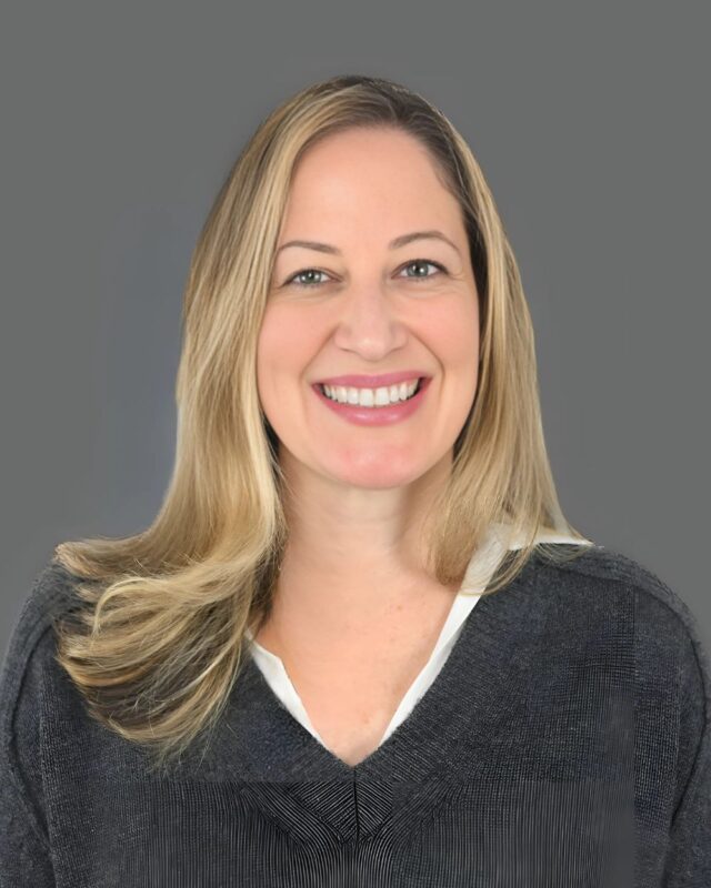 Smiling person with long blond hair wearing a gray blouse against a gray background.