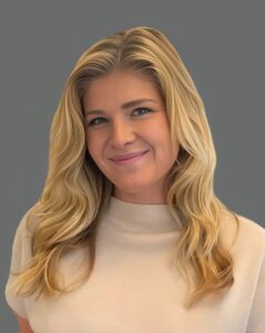 Smiling woman with long blonde hair wearing a light-colored sweater against a gray background.
