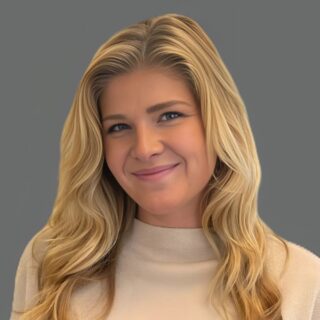 Smiling woman with long blonde hair wearing a light-colored sweater against a gray background.