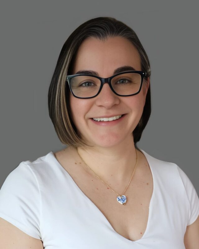 Person with glasses and short brown hair wearing a white shirt, smiling against a plain grey background.