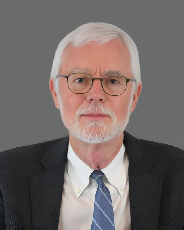 Elderly man with white hair and beard wearing glasses and a suit against a plain gray background.