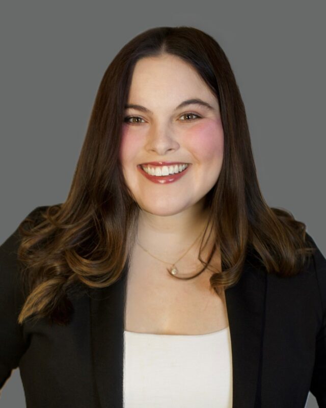 Smiling person with long dark hair wearing a black blazer and white top on a gray background