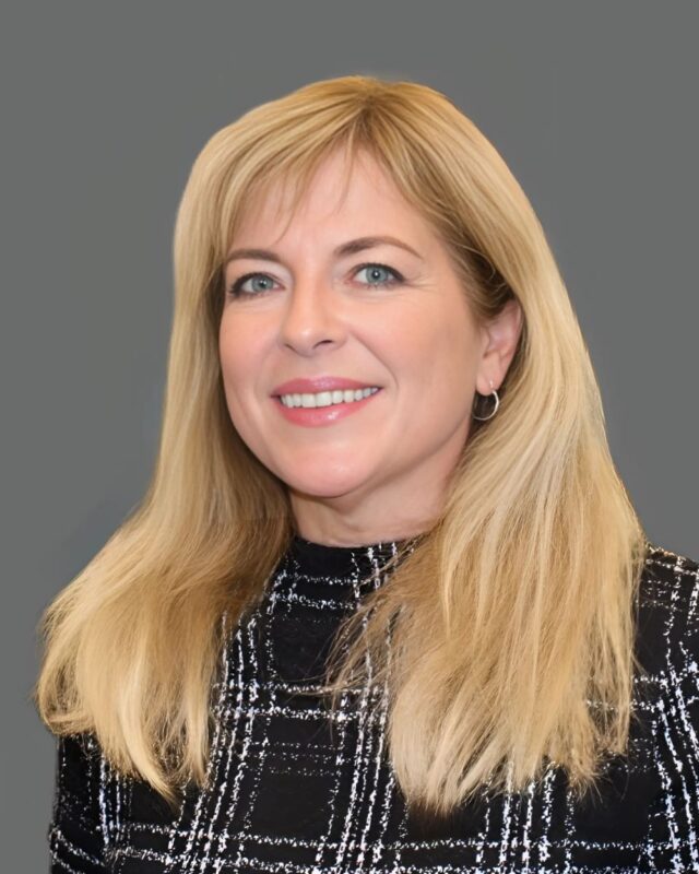 Smiling person with long blonde hair wearing a black patterned top against a gray background.