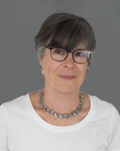 Woman wearing glasses and a silver necklace with short gray hair, dressed in a white top, against a gray background.