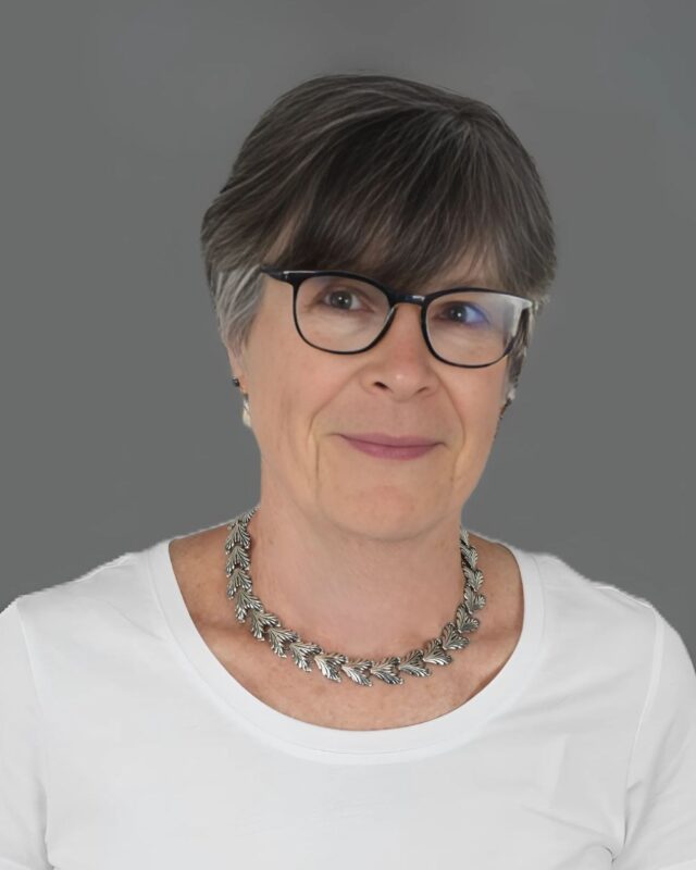 Woman wearing glasses and a silver necklace with short gray hair, dressed in a white top, against a gray background.