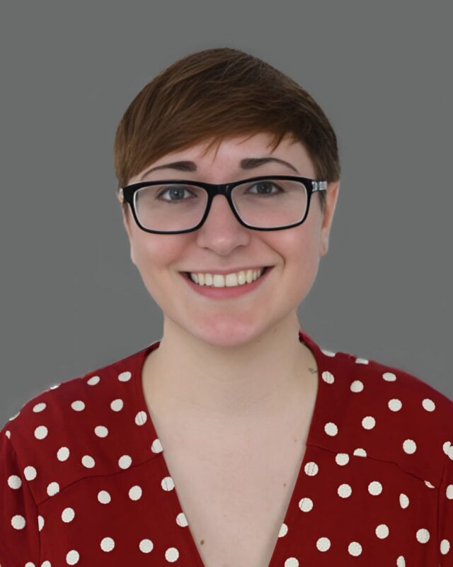 Person with short hair and glasses smiling, wearing a red polka dot shirt against a gray background.