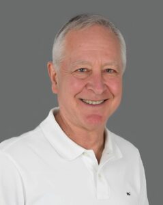 Smiling older man in a white polo shirt against a gray background.