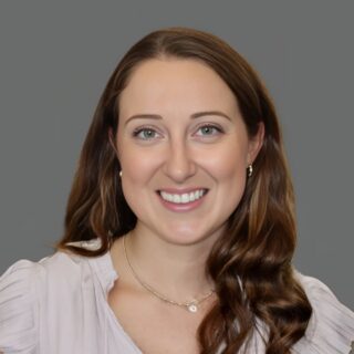 Smiling woman with long wavy brown hair wearing a light blouse, posing in front of a plain gray background.