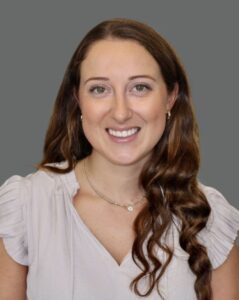 Portrait of a woman with long brown hair smiling, wearing a light blouse and necklace, set against a gray background.