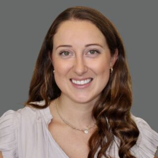 Portrait of a woman with long brown hair smiling, wearing a light blouse and necklace, set against a gray background.