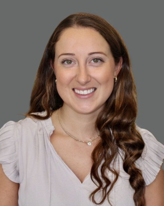Portrait of a woman with long brown hair smiling, wearing a light blouse and necklace, set against a gray background.