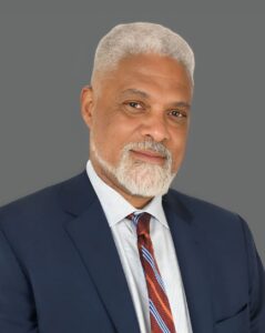 Man with a gray beard wearing a blue suit and striped tie against a gray background.