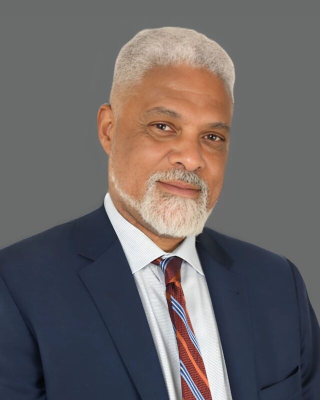 Man with a gray beard wearing a blue suit and striped tie against a gray background.