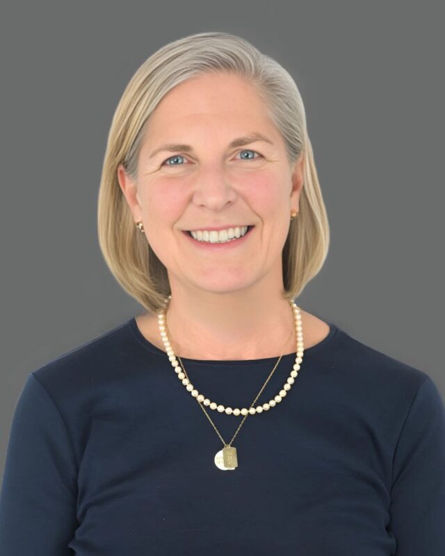 Smiling person with short blonde hair wearing a necklace and a navy top against a gray background.
