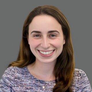 A person with long brown hair smiles at the camera while wearing a patterned top against a gray background.